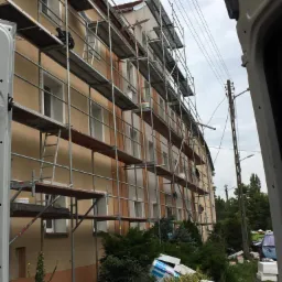 Budynek wielorodzinny w trakcie prac elewacyjnych, widoczne rusztowania, częściowo pomalowana fasada w odcieniach beżu i bieli, materiały budowlane i puste wiadra na pierwszym planie, pochmurne niebo.