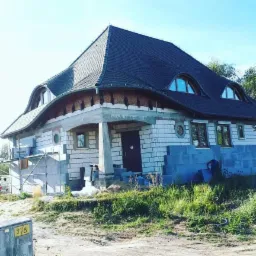Dom w trakcie budowy: murowane ściany z białej cegły silikatowej, nietypowy dach z lukarnami, widoczne bloczki betonowe i rusztowanie.