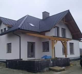 Dom jednorodzinny z jasną elewacją, ciemną dachówką i drewnianą konstrukcją nad wejściem, widok z poziomu gruntu, fragment czarnej skrzyni na pierwszym planie.