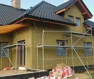 Dom w Wołowie w trakcie ocieplania elewacji, widoczne rusztowania i płyty izolacyjne mocowane do ściany. Częściowo ocieplona ściana w kolorze beżowym, widoczne punkty mocowania. Na pierwszym planie...