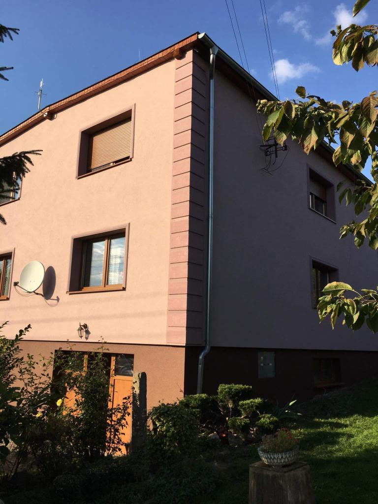 Dwukondygnacyjny dom z ocieploną elewacją w odcieniach beżu i brązu, z widocznymi detalami architektonicznymi w postaci boniowania narożników, brązowych ram okiennych i rolet zewnętrznych, antena...