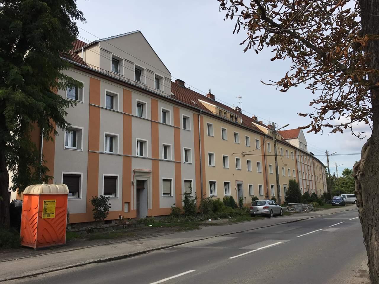 Długi budynek wielorodzinny z elewacją w odcieniach beżu, szarości i brązu, z pomarańczową przenośną toaletą na pierwszym planie i zaparkowanymi samochodami w tle.