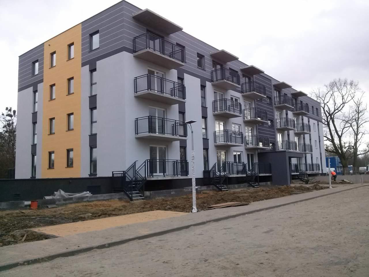 Nowoczesny budynek wielorodzinny z balkonami, wykończony w odcieniach szarości i żółci, widoczny fragment terenu przed budynkiem w trakcie prac wykończeniowych.