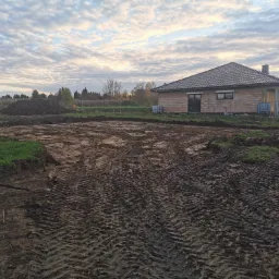 Przygotowany teren pod budowę domu jednorodzinnego, widoczne ślady opon maszyn budowlanych na ubitej ziemi, w tle budynek w stanie surowym otwartym i sterta ziemi, pochmurne niebo.