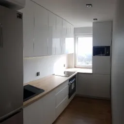 Nowoczesna kuchnia z białymi, lakierowanymi szafkami, drewnianym blatem, czarnym zlewem i płytą indukcyjną, oświetlona światłem dziennym wpadającym przez okno.