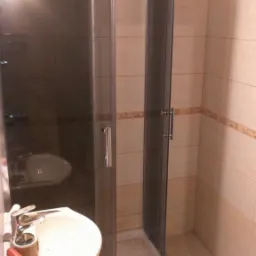 Narożna kabina prysznicowa z ciemnymi szybami, umywalka na postumencie z baterią, płytki beżowe z dekoracyjnym paskiem, widoczne narzędzia na umywalce.