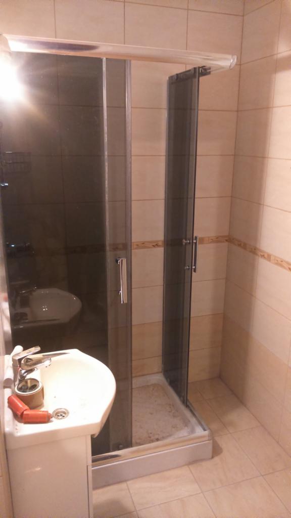 Narożna kabina prysznicowa z ciemnymi szybami, umywalka na postumencie z baterią, płytki beżowe z dekoracyjnym paskiem, widoczne narzędzia na umywalce.