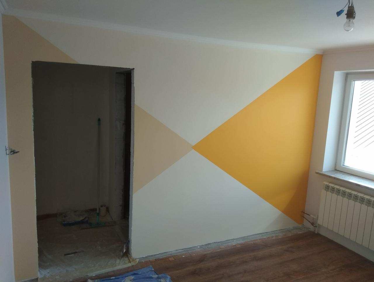 Pokój po malowaniu z geometrycznymi wzorami w odcieniach bieli, beżu i żółci. Widoczne niedokończone obramowanie drzwi i grzejnik pod oknem.