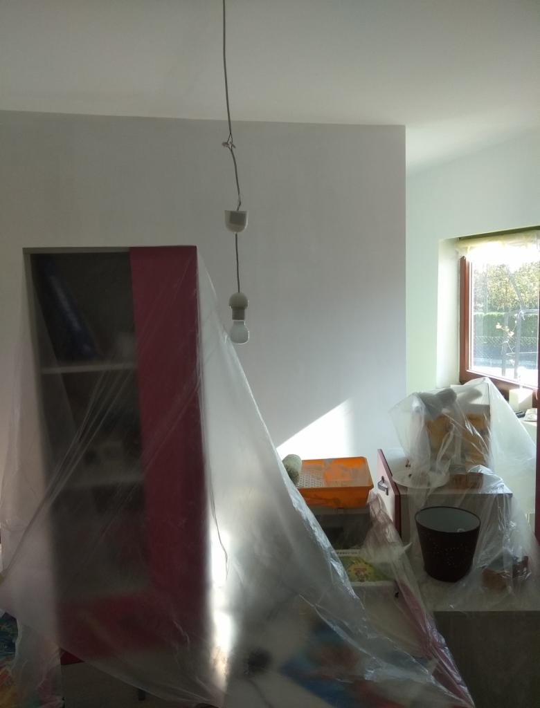 Pokój w trakcie przygotowań do malowania: meble zabezpieczone folią malarską, widoczne narzędzia malarskie i wisząca żarówka.