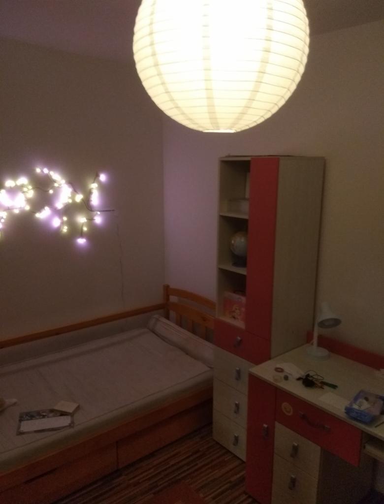 Pokój dziecięcy z drewnianym łóżkiem, biurkiem i regałem w kolorach bieli i czerwieni, oświetlony papierową kulą i girlandą lampek.