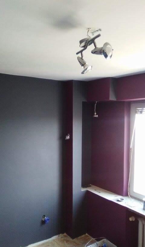 Pokój po malowaniu ścian na szaro i fioletowo, widoczna lampa sufitowa z trzema reflektorami, gniazdka elektryczne i grzejnik pod oknem.