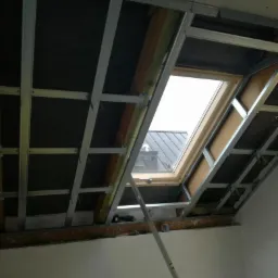 Szkielet metalowy pod zabudowę poddasza z widocznym oknem dachowym i częściowo ułożoną izolacją, widok z dołu na sufit poddasza, drabina oparta o ścianę.
