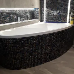 Biała wanna obudowana mozaiką w odcieniach brązu i granatu, z podświetleniem LED, chromowana bateria umywalkowa na tle szarej ściany i mozaiki.
