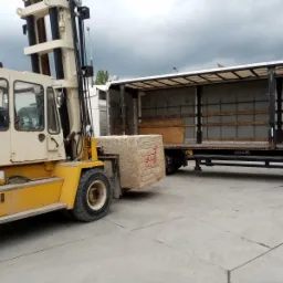 W Gdyni wózek widłowy z żółtymi elementami transportuje duży, szary blok kamienny z czerwonymi oznaczeniami do naczepy ciężarówki z otwartą plandeką.