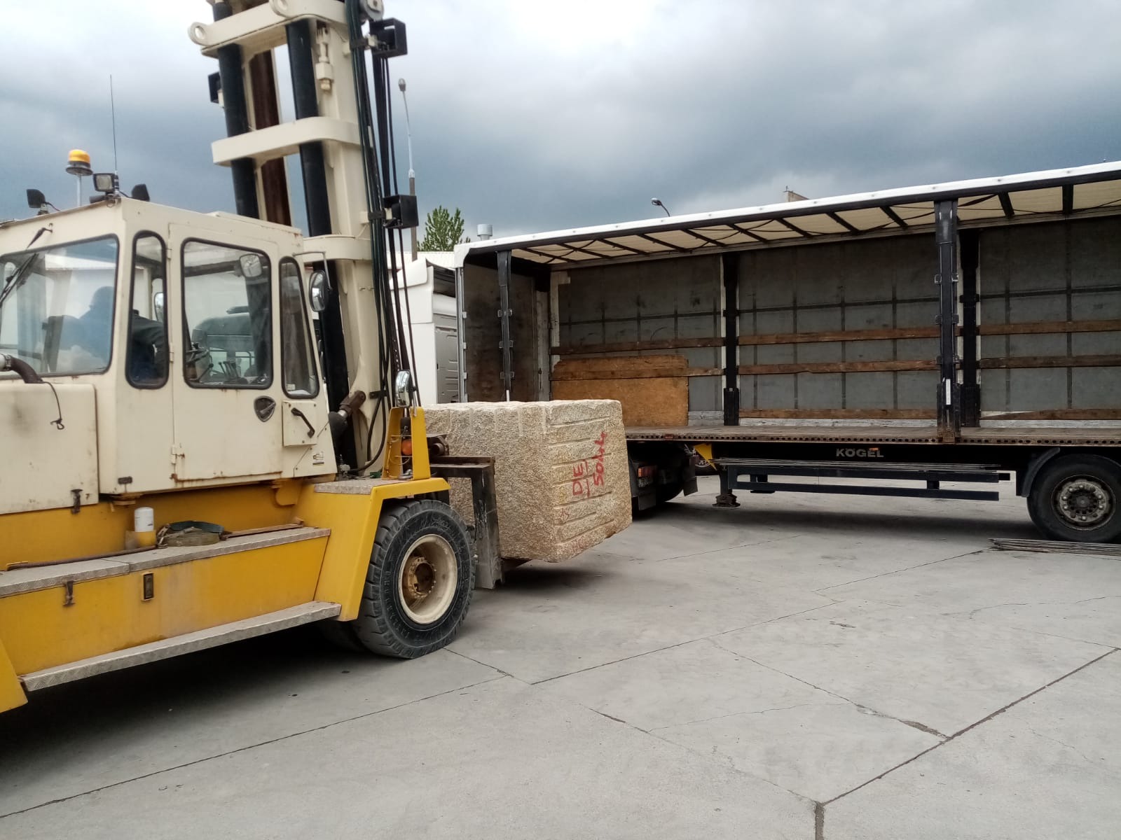 W Gdyni wózek widłowy z żółtymi elementami transportuje duży, szary blok kamienny z czerwonymi oznaczeniami do naczepy ciężarówki z otwartą plandeką.