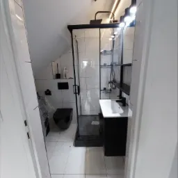 Nowoczesna łazienka na poddaszu z czarną armaturą, kabiną prysznicową typu walk-in, wiszącą toaletą i umywalką na szafce, oświetlona listwą LED.