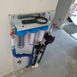 Zamontowany, potrójny filtr wody z niebieskimi obudowami, widoczne rury, zawory i manometr. Na górze klucz do odkręcania obudów filtrów. Podłoże surowe, w trakcie wykończenia.