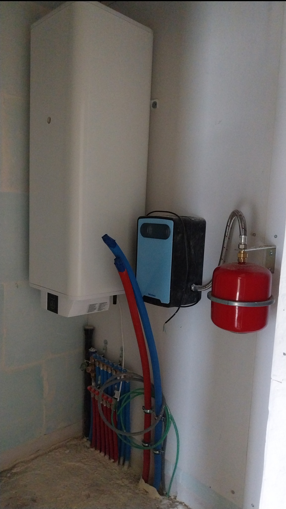 Nowa instalacja grzewcza z białym zasobnikiem, niebieskim modułem sterującym i czerwonym naczyniem przeponowym, widoczne rury w kolorach czerwonym i niebieskim oraz liczne złączki na ścianie...