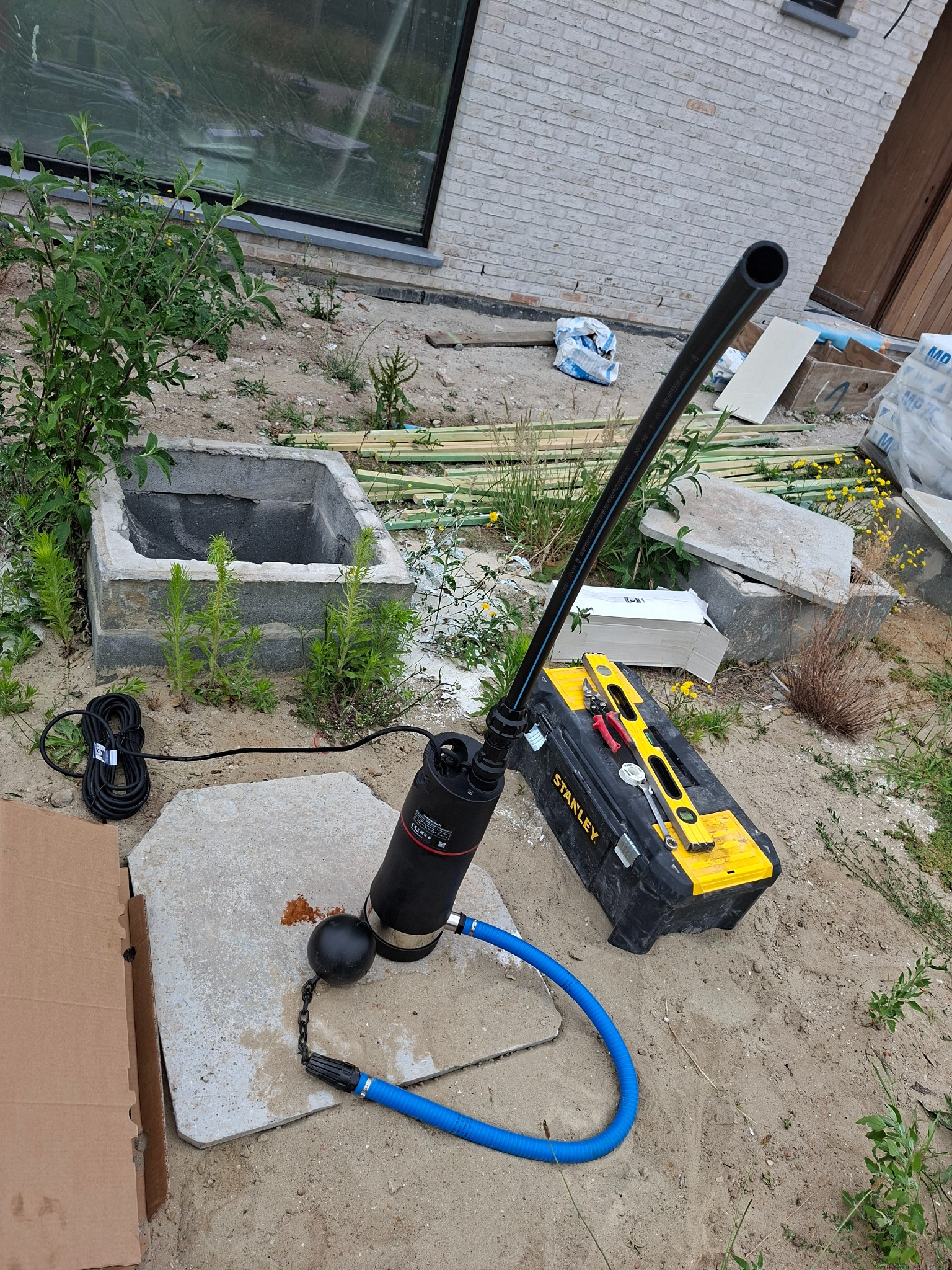 Pompa zanurzeniowa z czarnym pływakiem i niebieskim wężem, ustawiona obok betonowego zbiornika i skrzynki narzędziowej Stanley na piaszczystym podłożu.