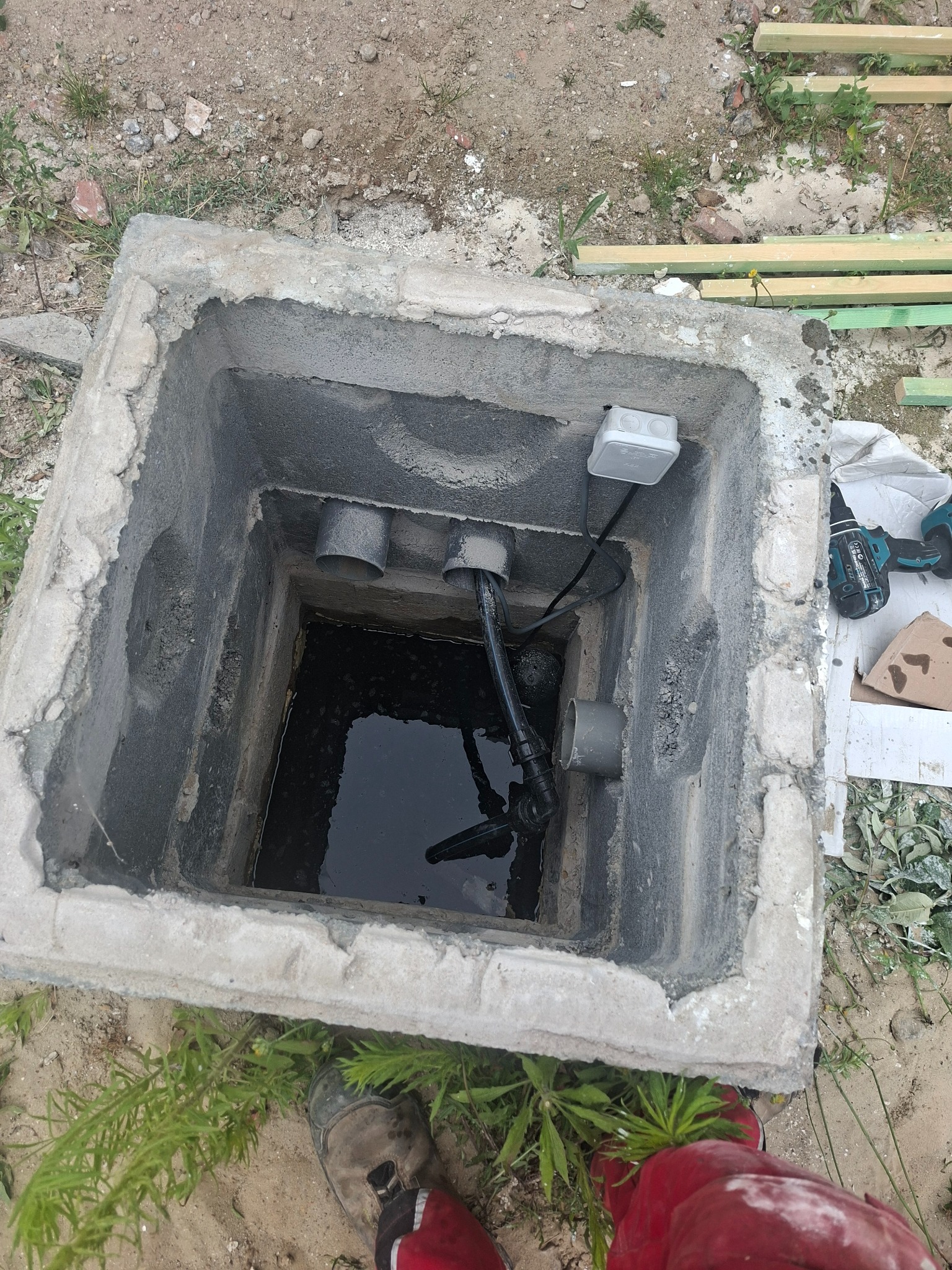Betonowy zbiornik z pompą zanurzoną w wodzie, widoczne rury i skrzynka elektryczna. W tle narzędzia i elementy drewniane, co sugeruje prace instalacyjne w toku.