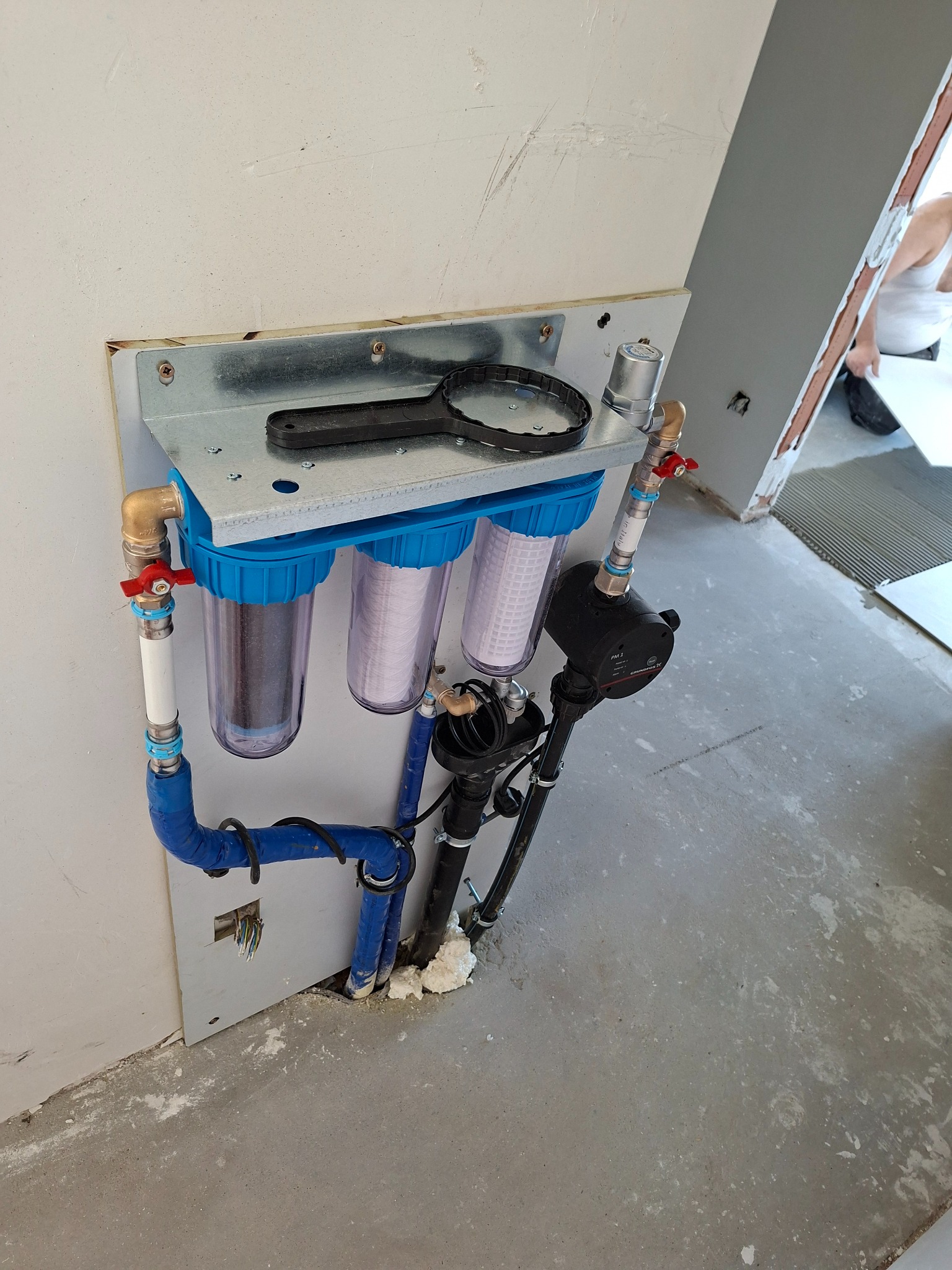 Zamontowany, potrójny filtr wody z niebieskimi obudowami, widoczne rury, zawory i manometr. Na górze klucz do odkręcania obudów filtrów. Podłoże surowe, w trakcie wykończenia.