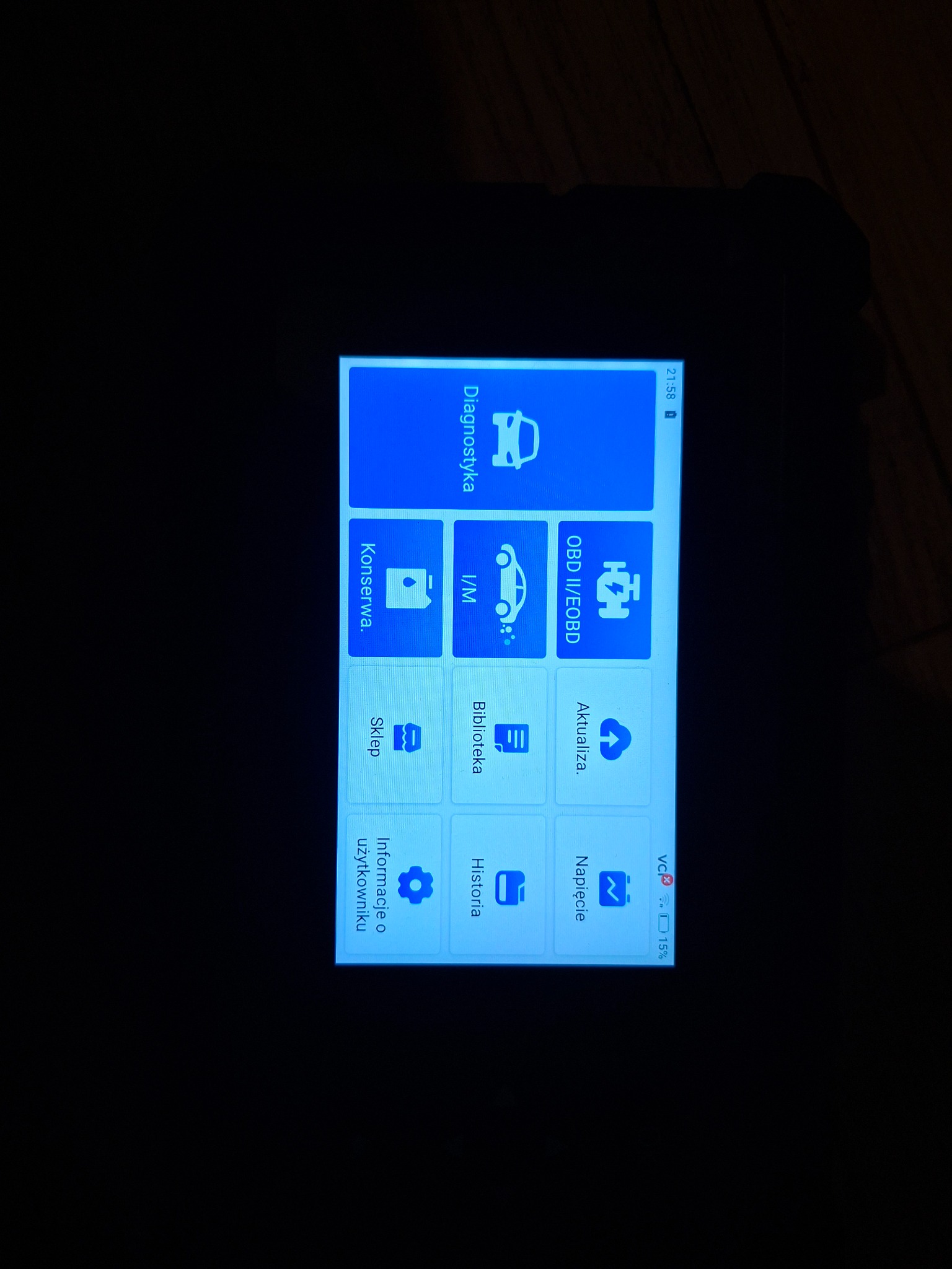 Ekran urządzenia diagnostycznego z ikonami: Diagnostyka, OBD II/EOBD, I/M, Konserwa, Aktualizacja, Biblioteka, Historia, Napięcie, Informacje o użytkowniku. Godzina 21:58, bateria 15%.