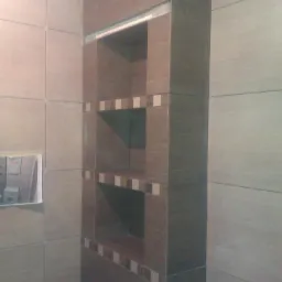 Wnęka z półkami wyłożona brązowymi płytkami w łazience, dekoracyjne akcenty z mozaiki, widoczna instalacja wentylacyjna.