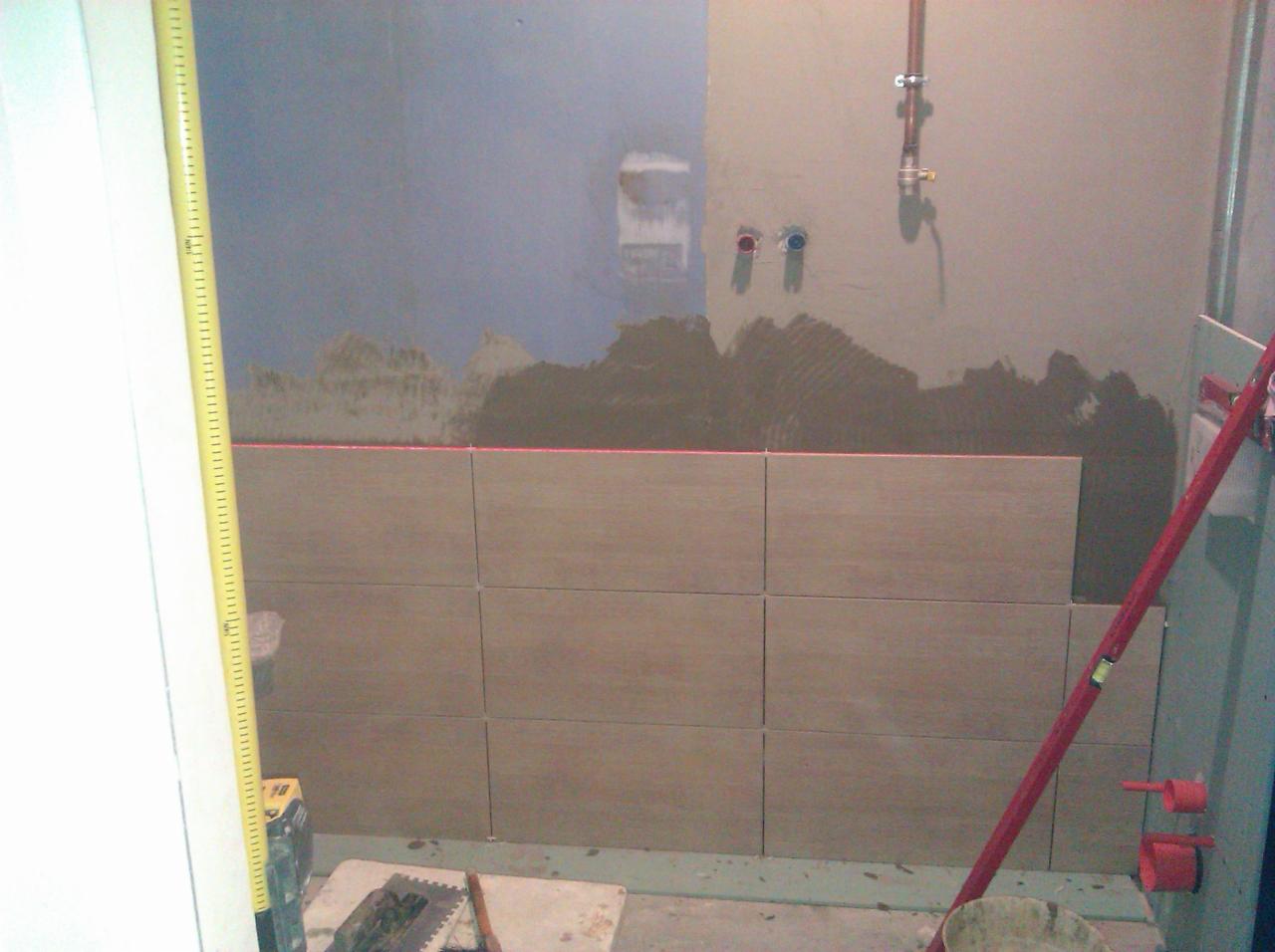 Ściana w trakcie układania płytek ceramicznych, widoczna poziomica laserowa, fragmenty ściany z nałożonym klejem i instalacją hydrauliczną