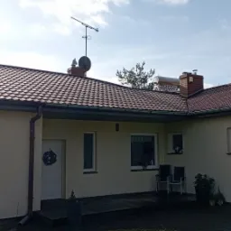 Widok z zewnątrz na parterowy dom z brązowym dachem z dachówki, beżową elewacją, ciemnymi rynnami i białymi oknami, na dachu widoczna antena i kolektory słoneczne, na drzwiach wejściowych wieniec.