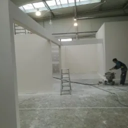 Wnętrze w trakcie prac wykończeniowych: widoczne ściany z płyt karton gips, aluminiowa drabina oraz pracownik używający szlifierki do gładzi, pylącej na posadzkę.