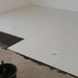 Wesley D Ogólnobudowlane - Białe płytki podłogowe, częściowo ułożone na zaprawie, widoczny kubeł z narzędziami, w tle fragment ściany i okna.