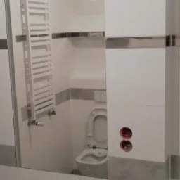 Wykończona łazienka z białymi płytkami, szarym pasem dekoracyjnym i chromowanymi listwami, widoczny grzejnik drabinkowy, toaleta i puszki elektryczne bez osprzętu.