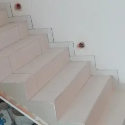 Wykończone płytkami schody wewnętrzne z widocznymi puszkami elektrycznymi w ścianie i workami zaprawy klejowej na podłodze.