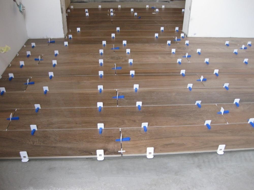 Układanie brązowych płytek podłogowych imitujących drewno z użyciem białych i niebieskich krzyżyków dystansowych, widoczne fugi i surowy beton podłoża.