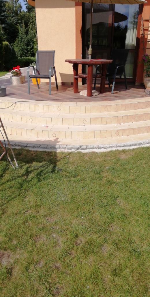 Wykończony taras z beżowych płytek z dekoracyjnymi wstawkami, schody z obrzeżem z kamienia, meble ogrodowe i parasol na tle domu z ogrodem.