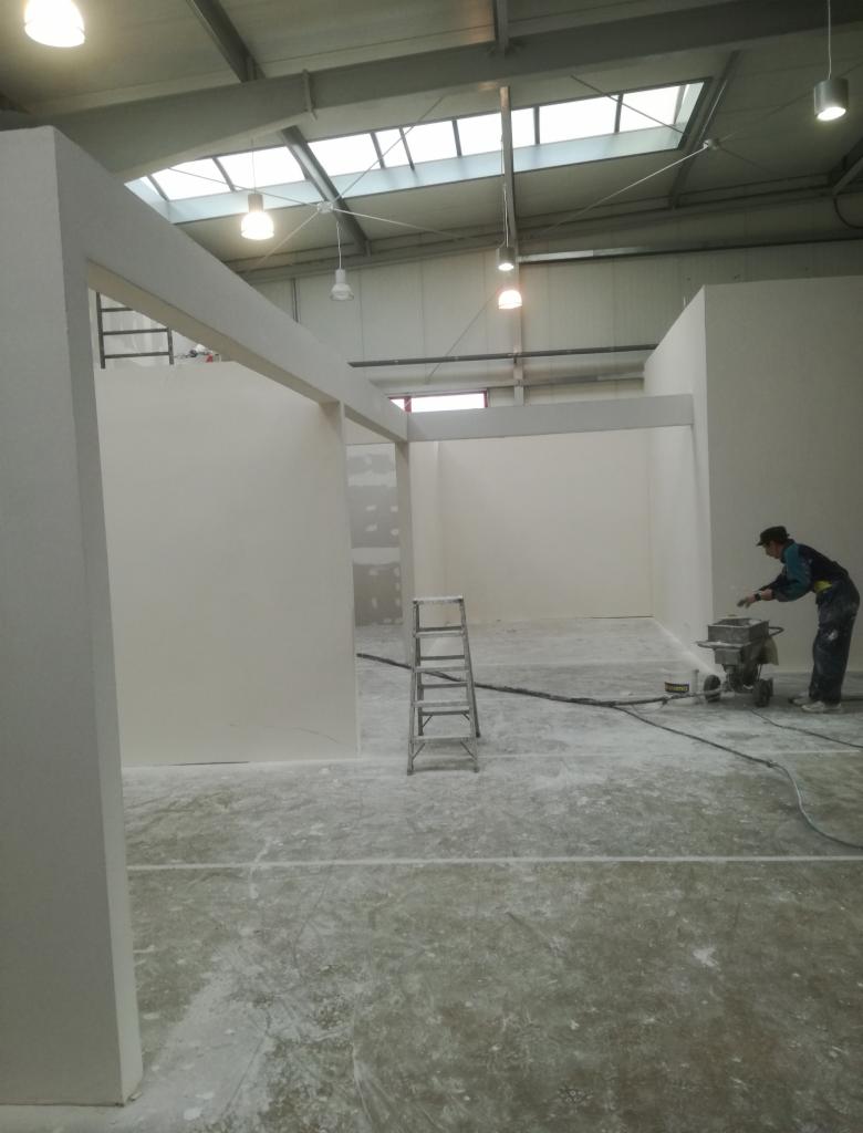 Wnętrze w trakcie prac wykończeniowych: widoczne ściany z płyt karton gips, aluminiowa drabina oraz pracownik używający szlifierki do gładzi, pylącej na posadzkę.