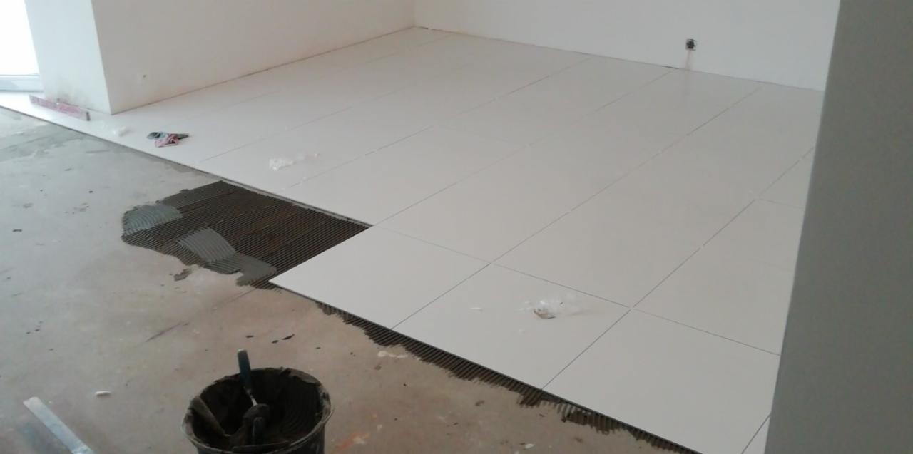 Białe płytki podłogowe, częściowo ułożone na zaprawie, widoczny kubeł z narzędziami, w tle fragment ściany i okna.