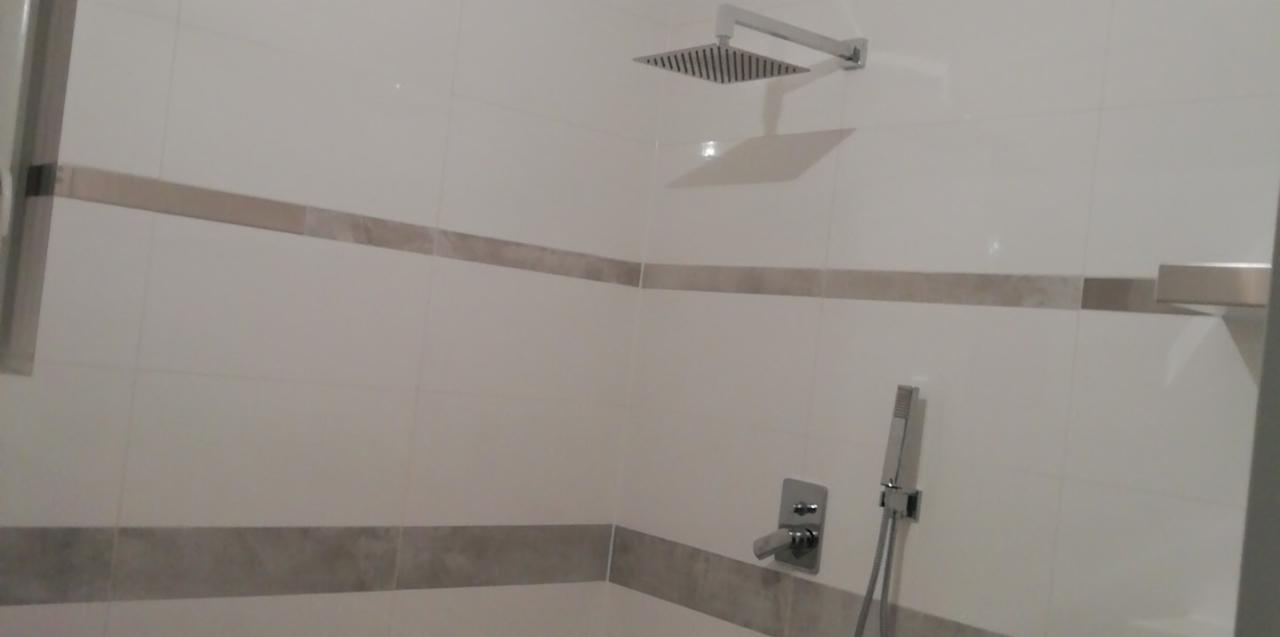 Wykończona kabina prysznicowa z białą glazurą i dekoracyjnym pasem z płytek w odcieniach szarości, widoczna deszczownica i bateria natryskowa.