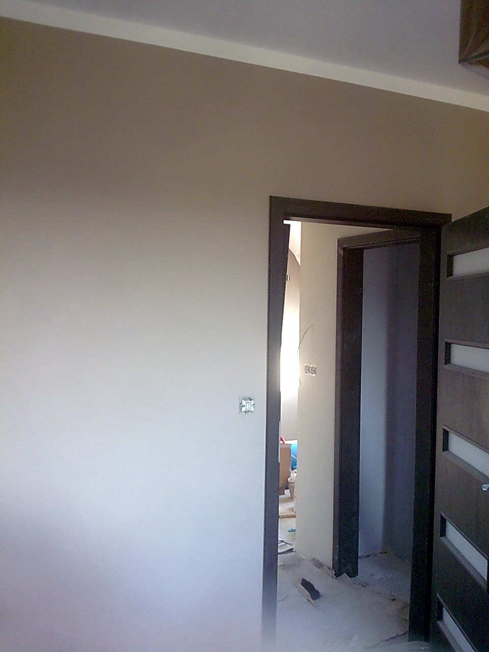 Świeżo pomalowana ściana w odcieniach beżu, z widocznym gniazdkiem elektrycznym, ciemna ościeżnica drzwiowa i fragment drzwi z przeszkleniami, w tle widoczne przygotowania do dalszych prac...