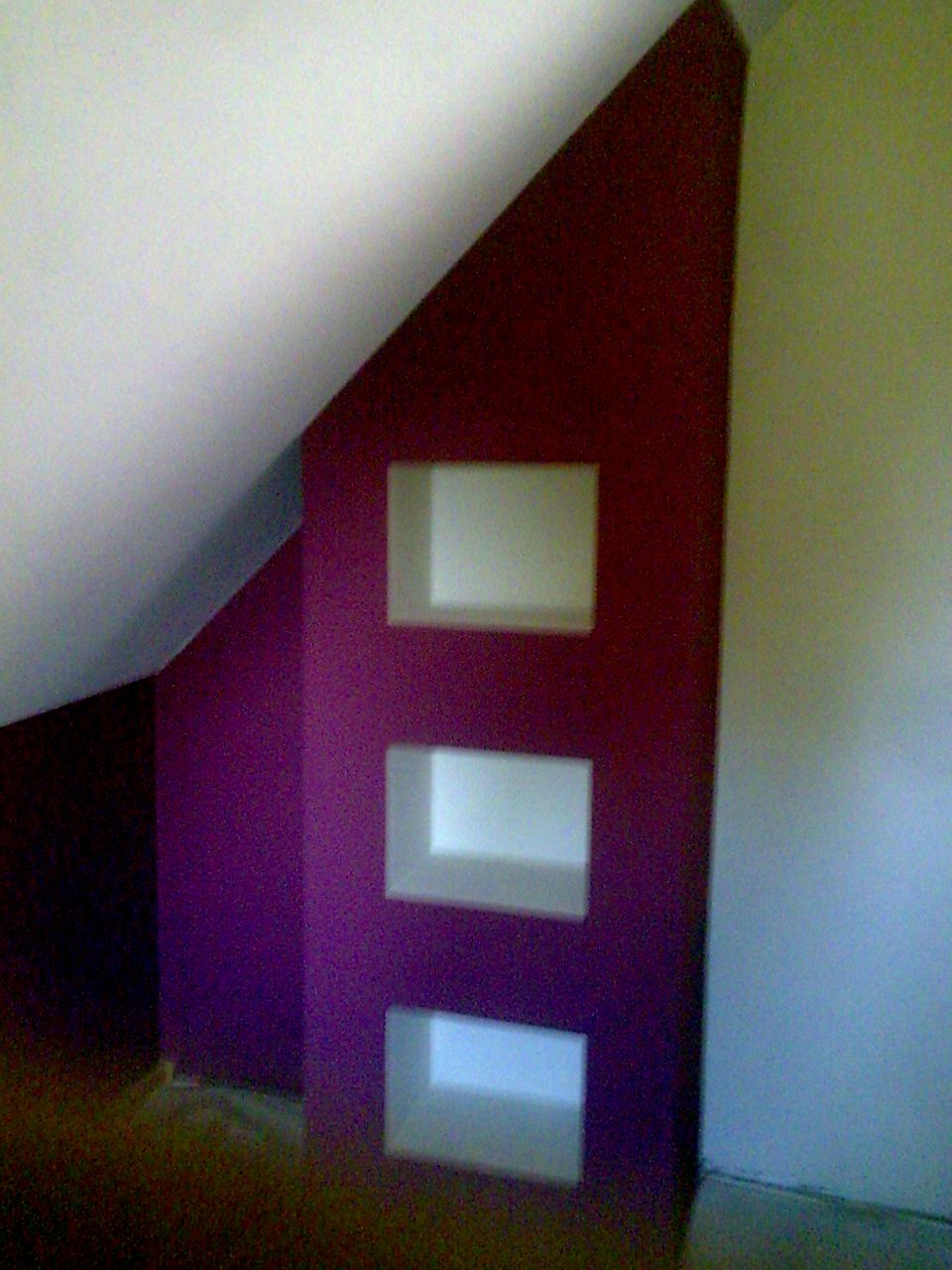 Wnęka w ścianie pod skosem dachu, pomalowana na fioletowo-bordowo, z trzema prostokątnymi, białymi półkami.