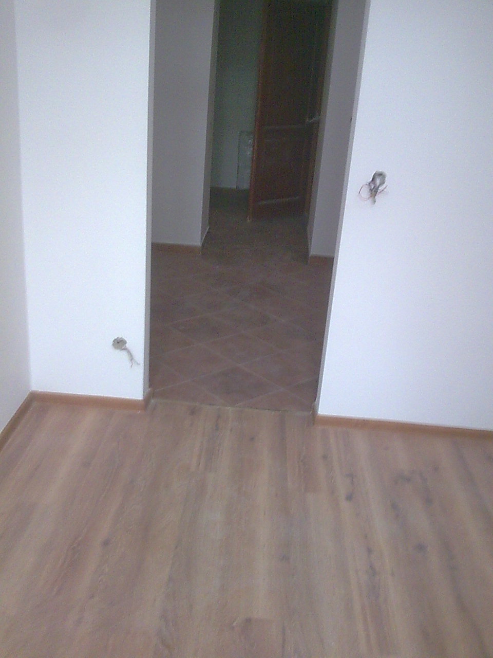 Fragment pokoju z ułożoną podłogą z paneli w odcieniach drewna i przejściem do innego pomieszczenia z brązową, ułożoną w karo podłogą z płytek. Widoczne wystające kable ze ściany.