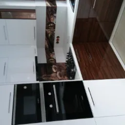 Nowoczesna kuchnia z białymi i brązowymi szafkami, płyta gazowa i czarnymi piekarnikami w zabudowie, dekoracyjny panel nad płytą.