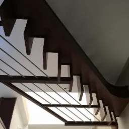 Nowoczesne schody z drewnianymi stopniami i metalowymi barierkami, widziane z dołu pod kątem, z widocznym fragmentem sufitu i ściany.