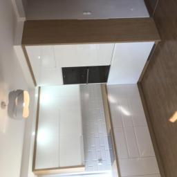 HAŁYS-Meble - Nowoczesna kuchnia z białymi, błyszczącymi frontami szafek i drewnianym blatem, oświetlona designerskim kinkietem na ścianie, z widoczną zabudowaną płytą grzewczą i białymi płytkami na ścianie.