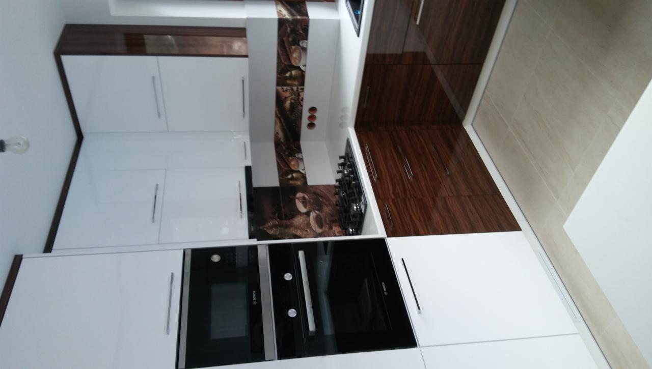 Nowoczesna kuchnia z białymi i brązowymi szafkami, płyta gazowa i czarnymi piekarnikami w zabudowie, dekoracyjny panel nad płytą.