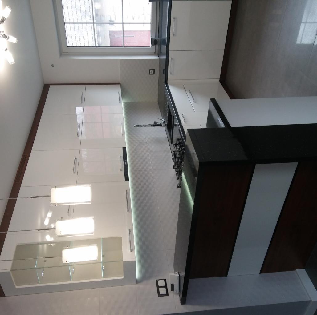 Nowoczesna, biała kuchnia z połyskiem, z czarnym blatem i barkiem z elementami drewna, oświetlenie LED pod szafkami i wiszące lampy, widok na okno.