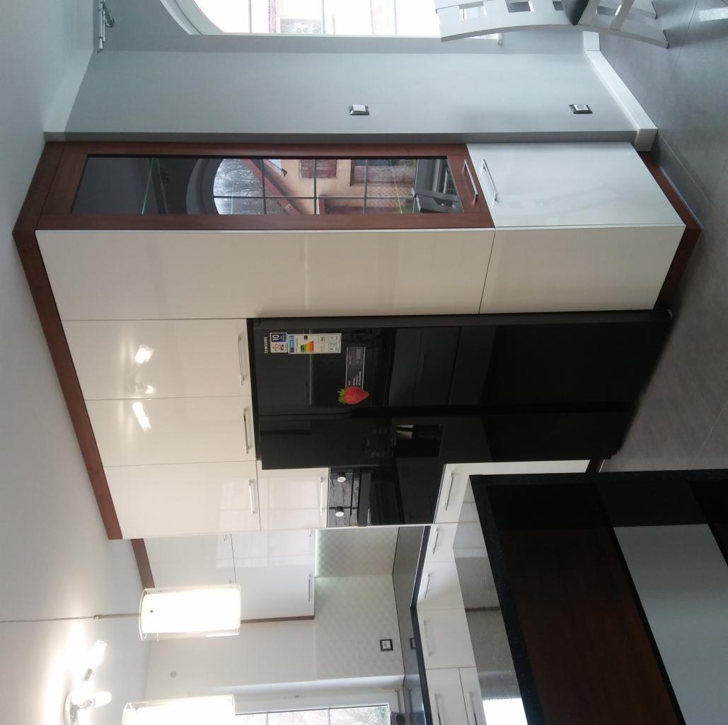 Nowoczesna kuchnia z białymi, lakierowanymi szafkami i czarną lodówką, z blatem w kolorze ciemnego drewna. Szafka wisząca z przeszklonym frontem i ciemną ramą. Widoczne elementy wyposażenia kuchni...