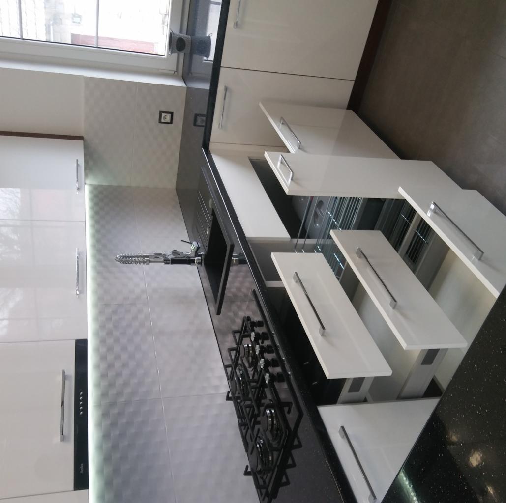 Nowoczesna, biała kuchnia z czarnym blatem i otwartymi szufladami, widok na płytę gazową i zlewozmywak, oświetlenie LED pod szafkami.