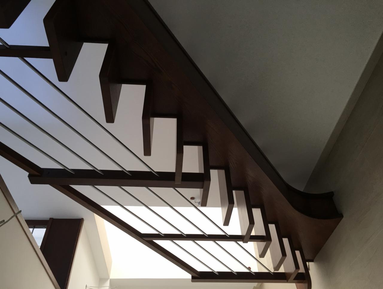 Nowoczesne schody z drewnianymi stopniami i metalowymi barierkami, widziane z dołu pod kątem, z widocznym fragmentem sufitu i ściany.