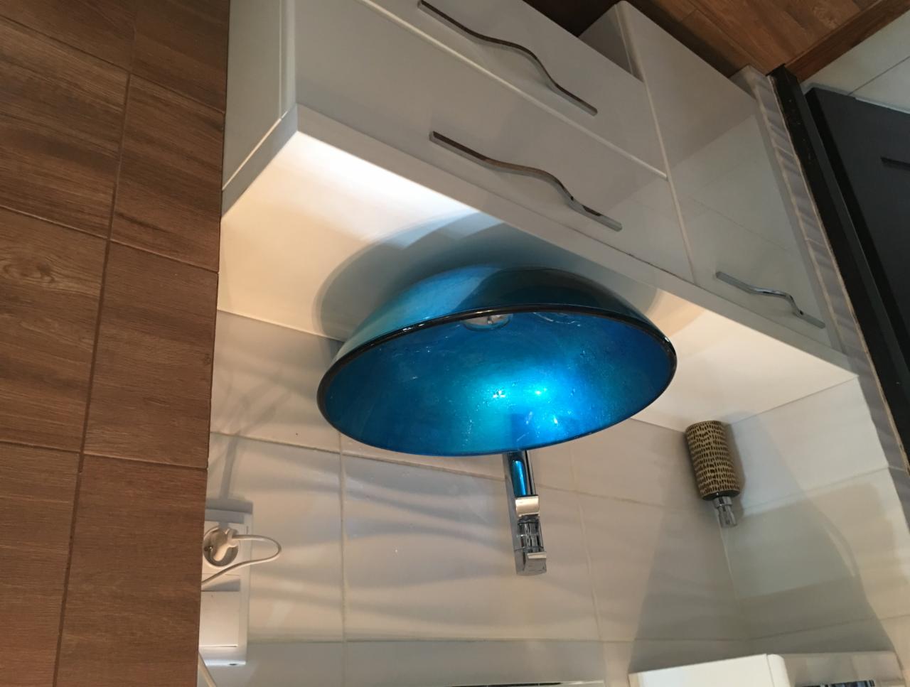 Błękitna szklana umywalka nablatowa z chromowaną baterią, zamontowana na białym blacie pod wiszącymi szafkami z metalowymi uchwytami, widok na ścianę wyłożoną płytkami imitującymi drewno.